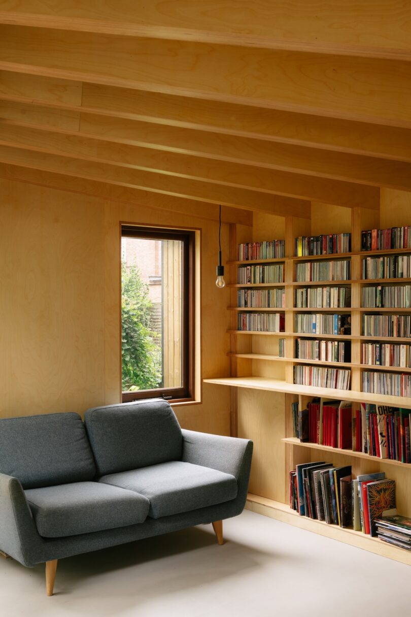 Room with a gray sofa and a wall-mounted bookshelf filled with books and CDs, next to a window.