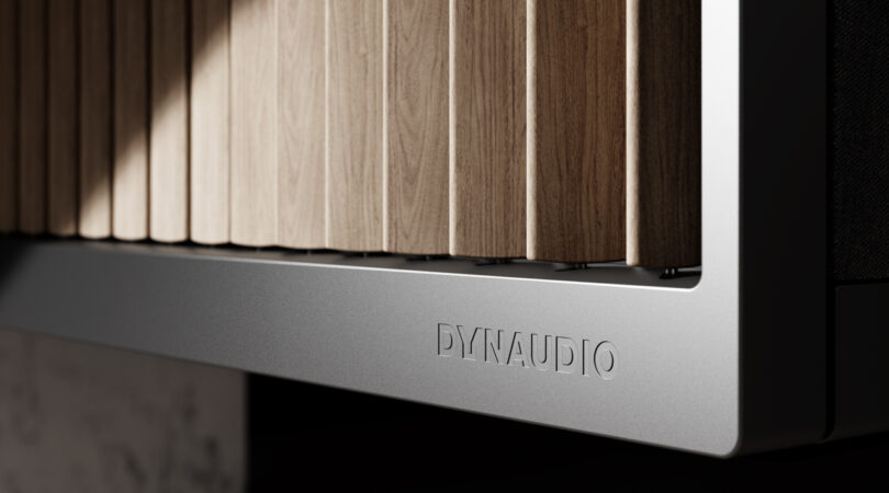 The close-up reveals a sleek speaker with vertical wooden slats and a metal edge, prominently showcasing the Dynaudio logo.