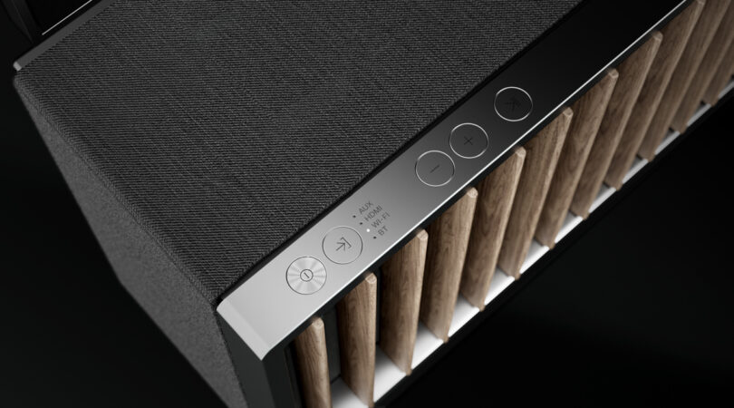 Close-up of a sleek Dynaudio speaker featuring a black textured top, four control buttons, and a vertical wooden slat design on the side.