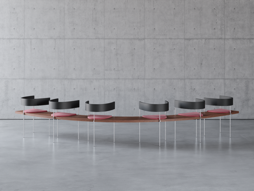 Modern curved wooden table with eight sleek black and pink chairs, set in a minimalistic room with a concrete wall and floor