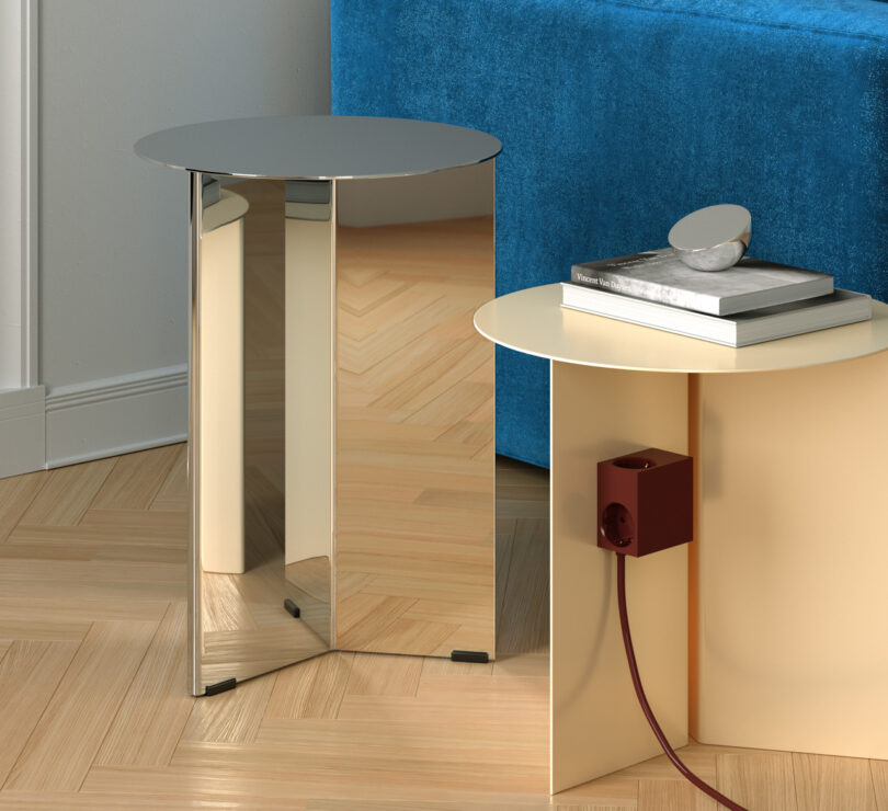 Two modern tables with reflective surfaces adorn the wooden floor. One table hosts books and a decorative object, while the other features a red electrical socket and cord, showcasing Kord's unique design flair.