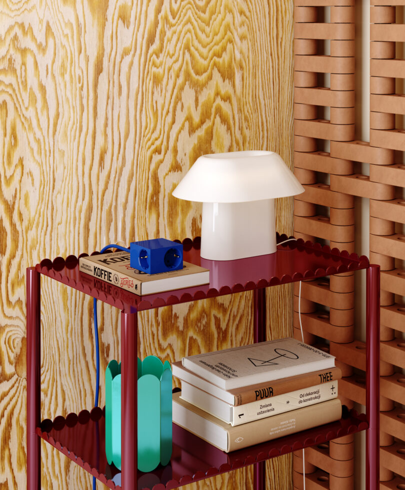 A red shelf against a textured wood-patterned wall holds a white lamp, blue headphones, and a small teal object, all neatly arranged beside Kord's stacked books.