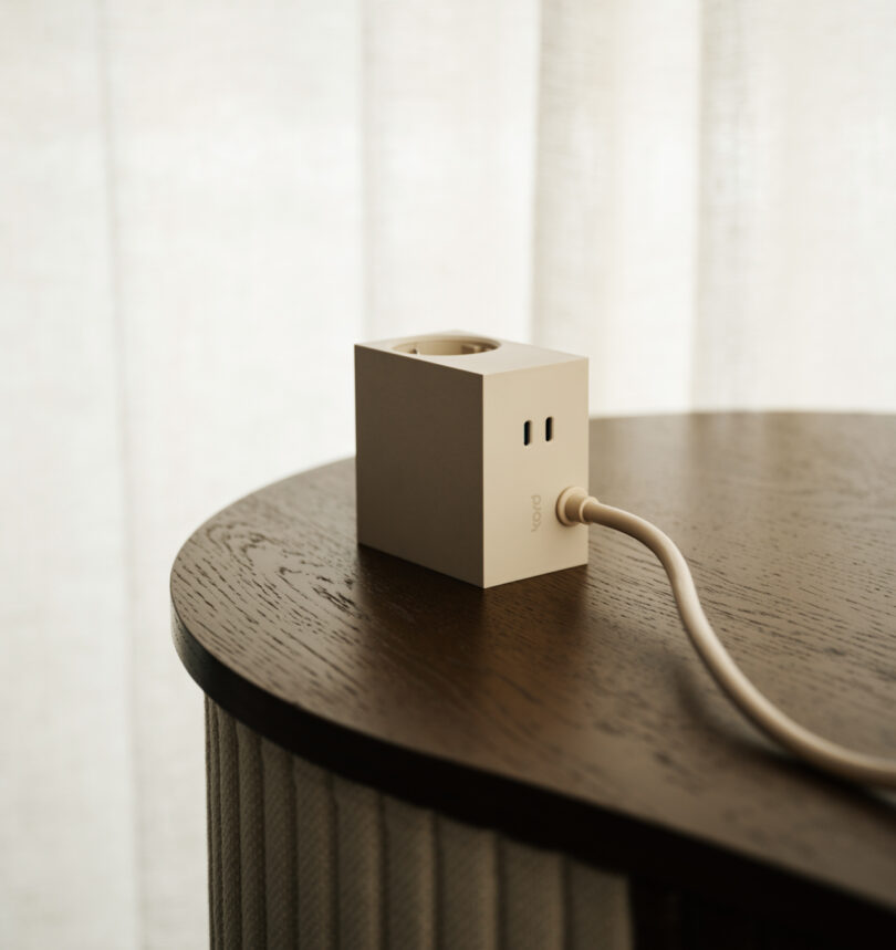 A white cube-shaped power adapter with a Kord plugged in rests on a wooden table, framed by light-colored curtains.