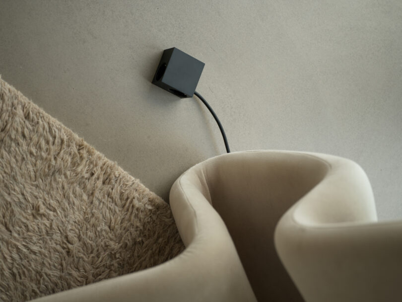A beige, curved chair sits beside a fluffy rug on a light gray floor, with a sleek black electronic device—Kord—subtly attached to a wire.