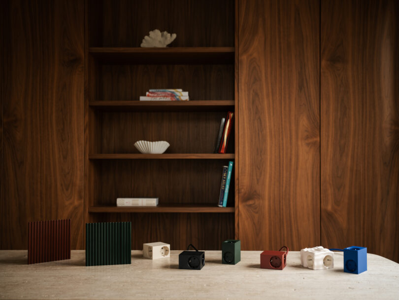 Colorful, cube-shaped Kord speakers and two ridged panels are displayed on a wooden table in front of a bookshelf with scattered books and decorative items.