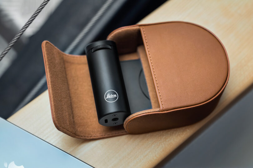 A black cylindrical Leica device with a Lux Grip feature nestles inside an open brown leather case, resting elegantly on a wooden surface.