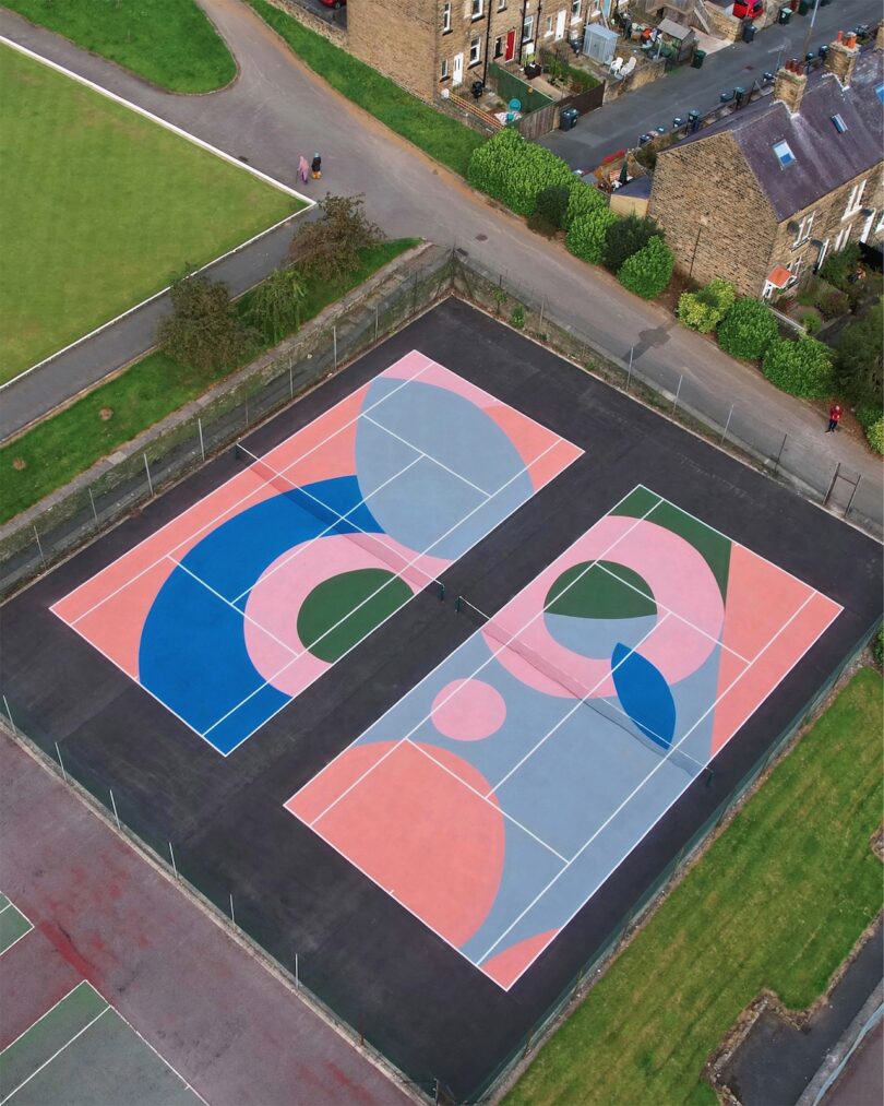 Aerial view of two tennis courts with colorful geometric designs, surrounded by a fence, adjacent to a grassy area and a road with houses