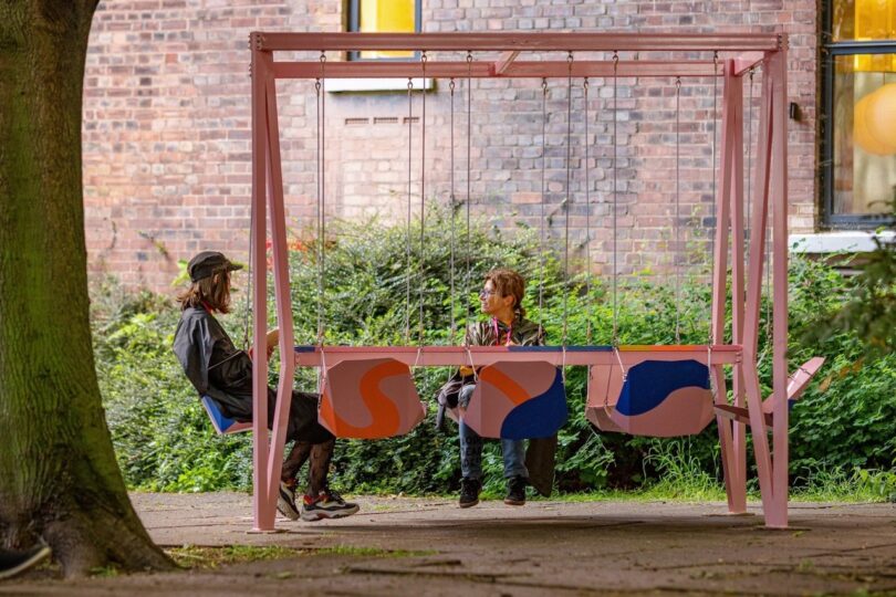 Two people sitting on a multi-seat pink swing set in a park, surrounded by greenery and a brick building in the background