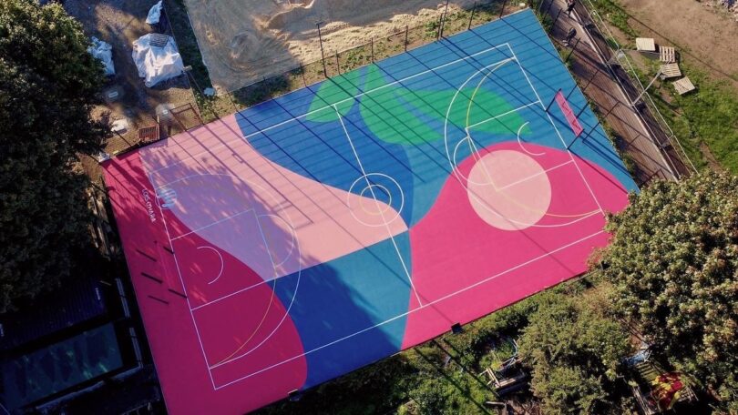 Aerial view of a colorful outdoor basketball court with abstract geometric shapes in blue, green, pink, and beige. Surrounded by trees and construction materials