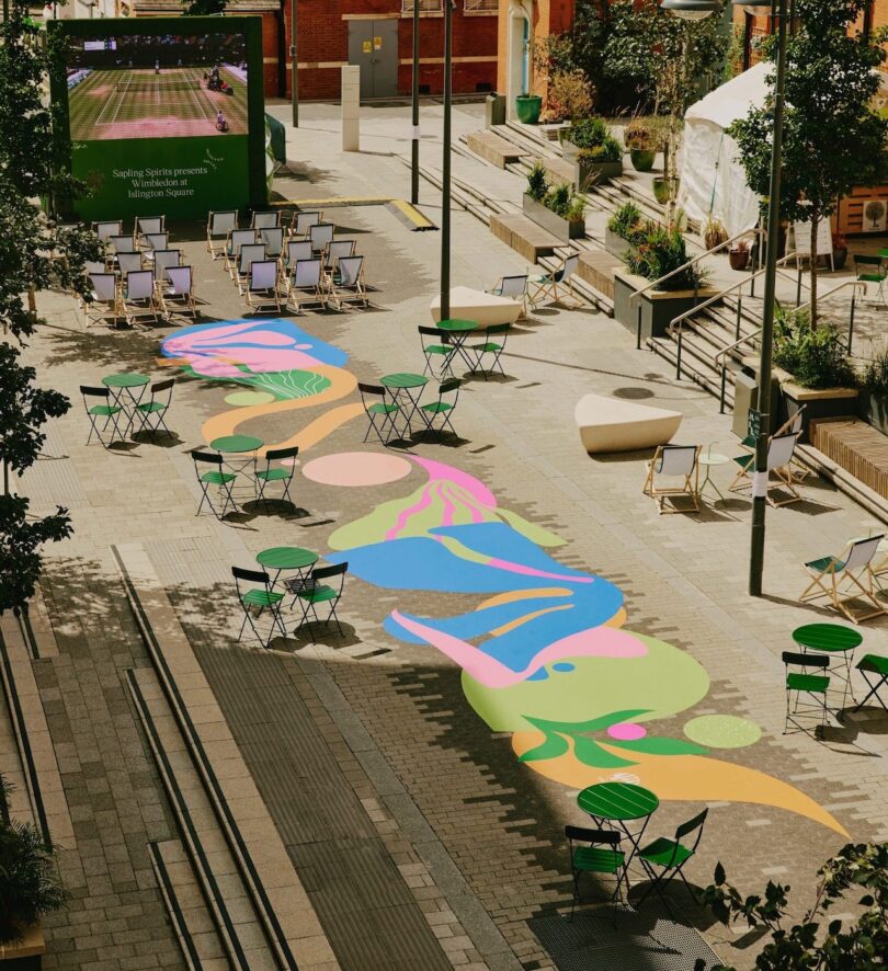 Outdoor seating area with colorful abstract patterns on the ground, green tables and chairs, and a screen displaying a sports game