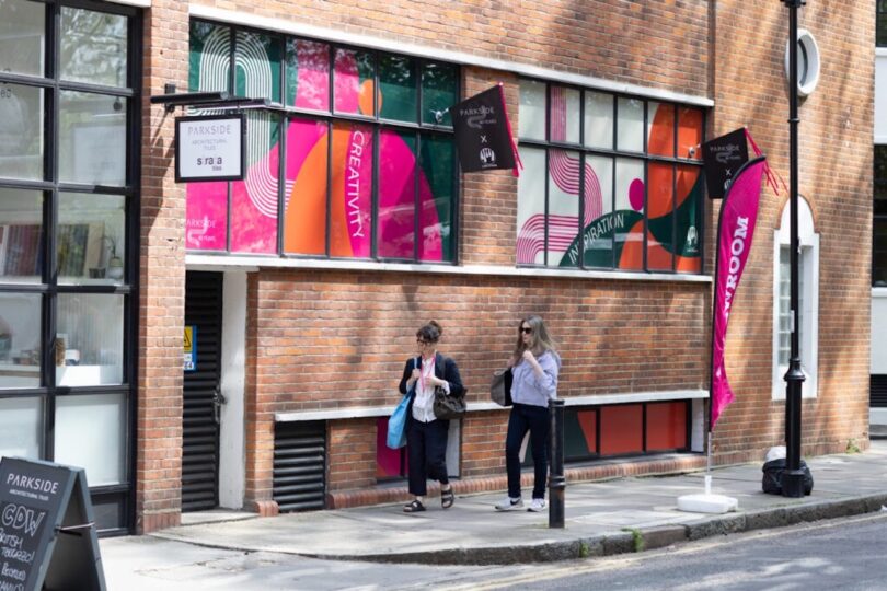 Two people walk past a brick building with colorful window decals and a "Parkside" sign. A pink flag displays words related to creativity and innovation