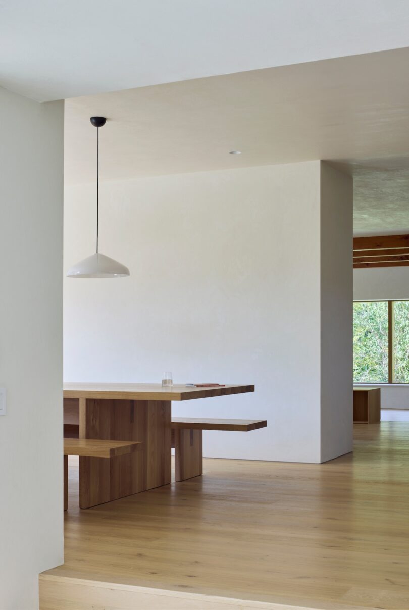 Minimalist dining area with a wooden table and benches, a white pendant light, light wood floors, and a large window showing greenery.