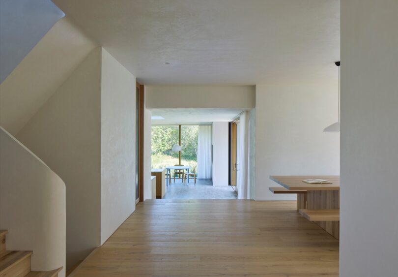 Minimalist interior with wooden floors, neutral walls, and large windows. A staircase on the left and a table with chairs visible in the distance. Bright, natural light fills the space.