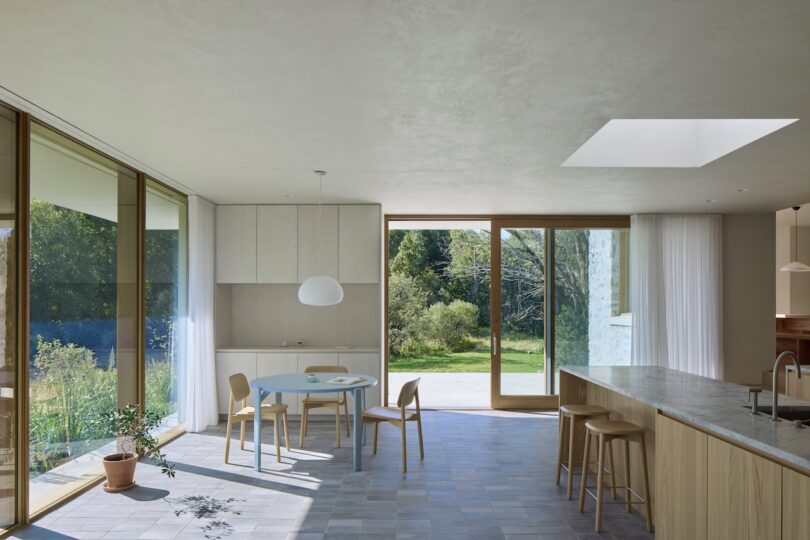 Modern living space with a glass wall overlooking a garden, featuring a round table, chairs, a small plant, and a kitchen island. Large windows and a skylight provide natural light.