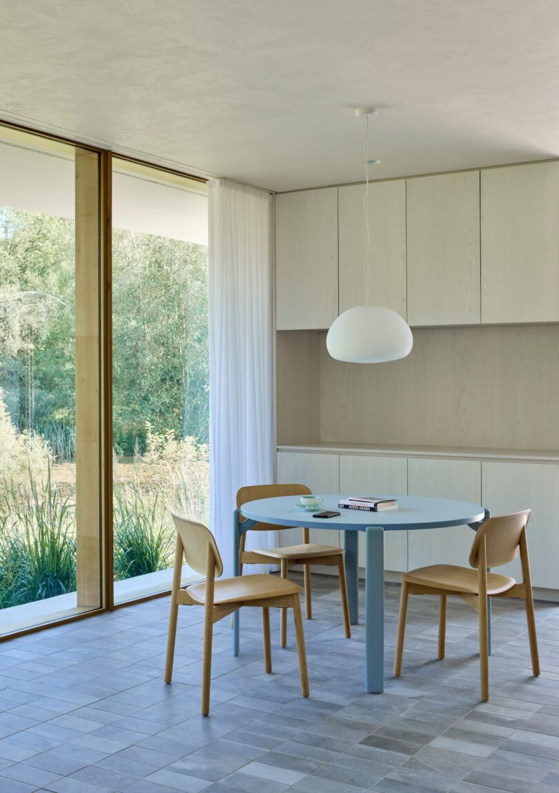 Minimalist dining area with a round blue table, three wooden chairs, and a hanging lamp. Large window reveals a garden view with trees and grass. Neutral color palette.