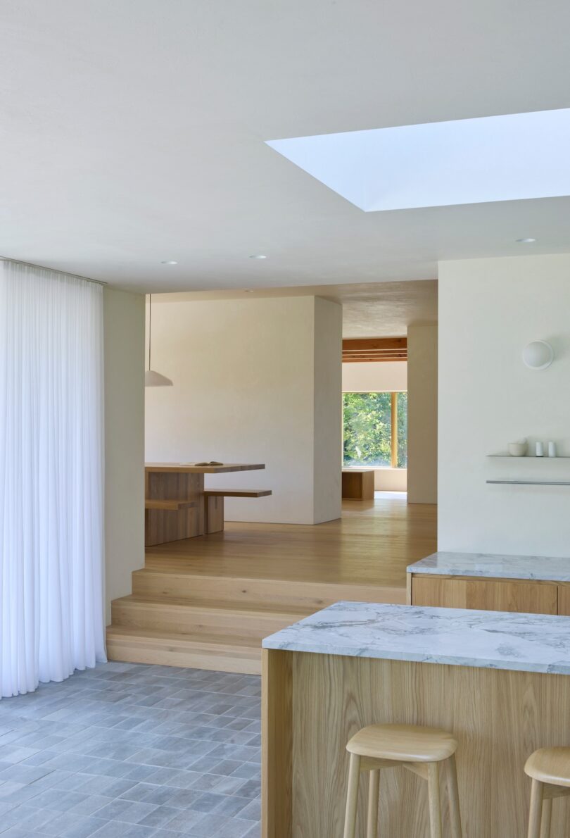 Minimalist open-plan interior with light wood flooring and furniture, a skylight, white walls, a kitchen island, and textured curtains in a bright, airy space.