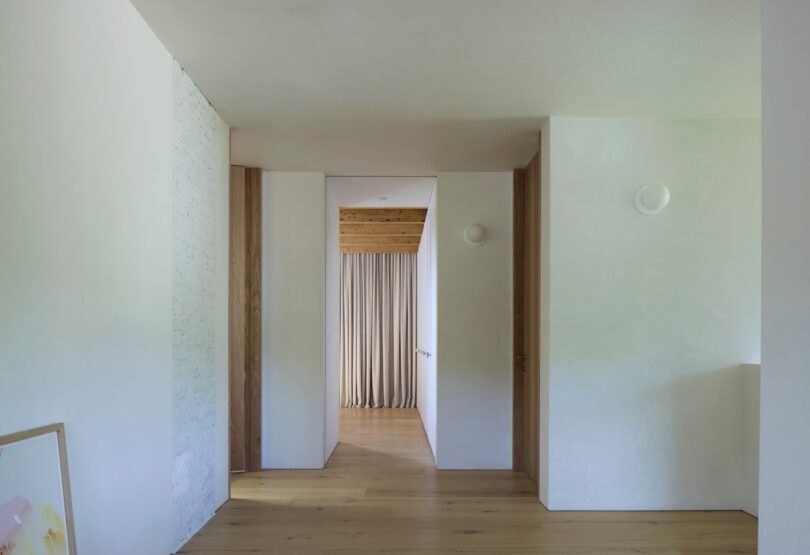 Minimalist hallway with wooden floors, white walls, and a doorway leading to a room with beige curtains and a wooden ceiling. A framed picture leans against the left wall.