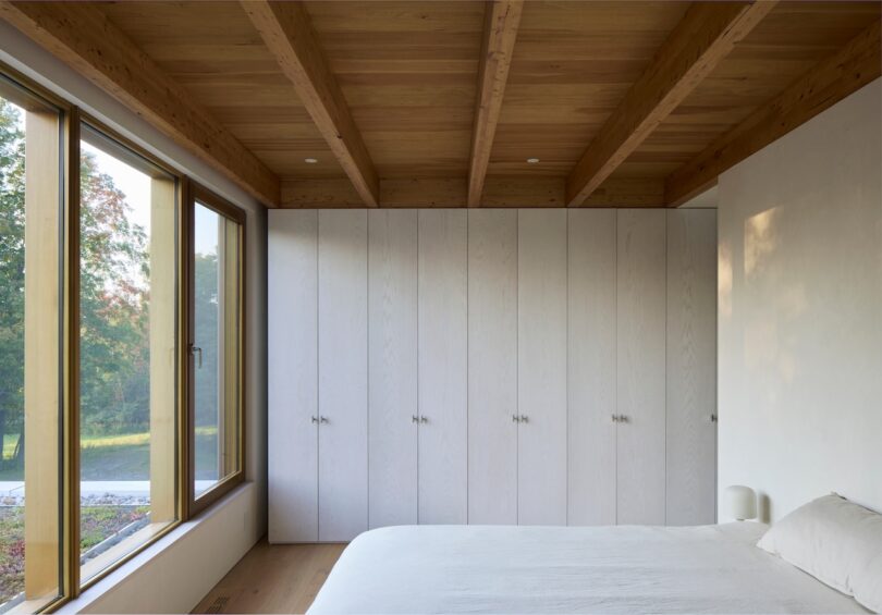 A minimalist bedroom with wooden beams, large windows, white walls, and built-in cabinets. A bed with white linens is in the foreground.