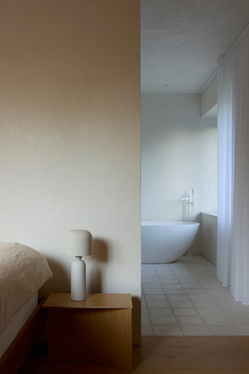 Minimalist bedroom with a wooden side table and white lamp. Adjacent bathroom features a freestanding bathtub and sheer curtain. Neutral tones dominate the space.