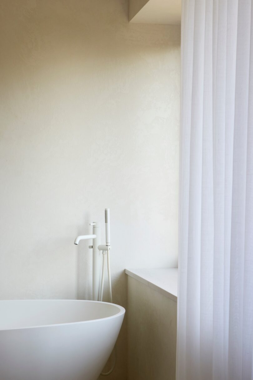 Minimalist bathroom with a white freestanding bathtub, handheld showerhead, and light beige walls. A sheer white curtain hangs beside the tub.