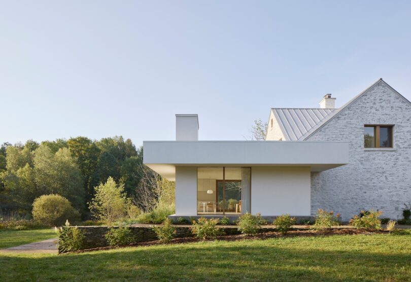 A modern house with a white facade and stone wall, featuring large windows and surrounded by greenery under a clear sky.