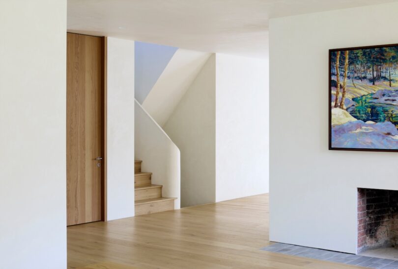 Minimalist interior with light wooden flooring, white walls, a wooden staircase, and a framed landscape painting above a brick fireplace.