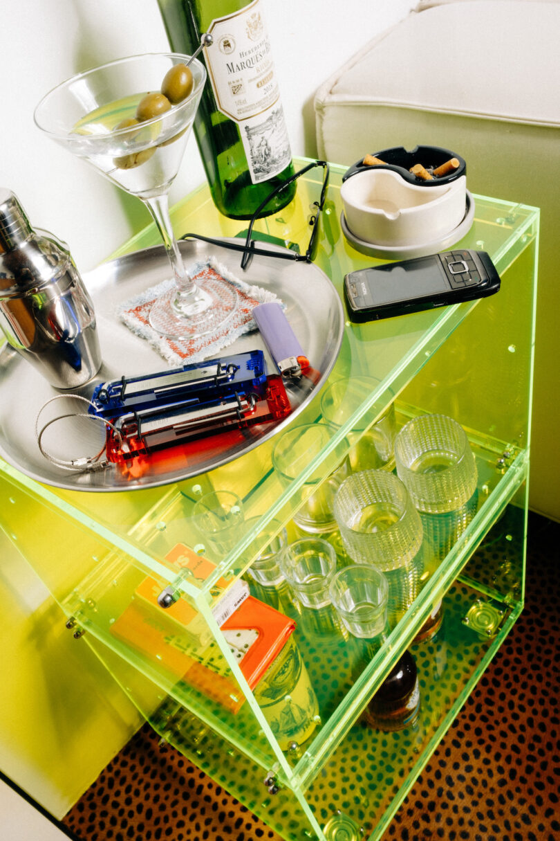 A glass side table with a martini, liquor bottle, lighter, cigarettes, glasses, and a cell phone on a tray. Below are additional glasses and a bottle