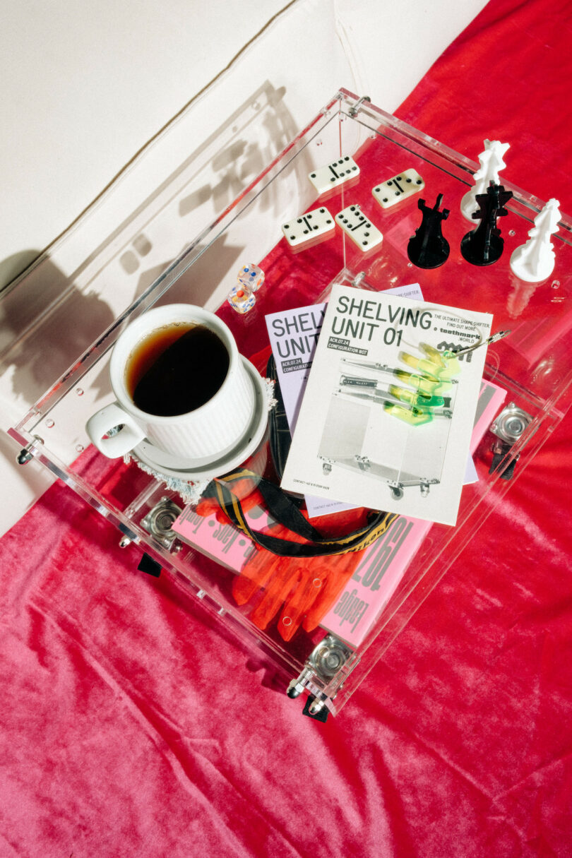 A table with a coffee cup, books, dominoes, chess pieces, and tools on a translucent container, set against a red velvet surface