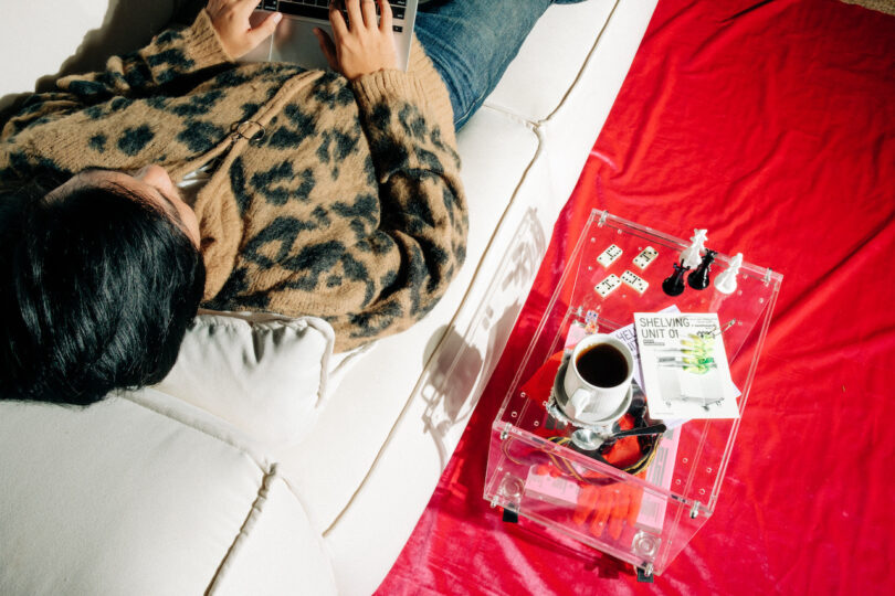 Person on a white couch typing on a laptop, wearing a leopard print sweater. A clear table nearby holds a coffee cup, a book, and playing cards on a red cloth