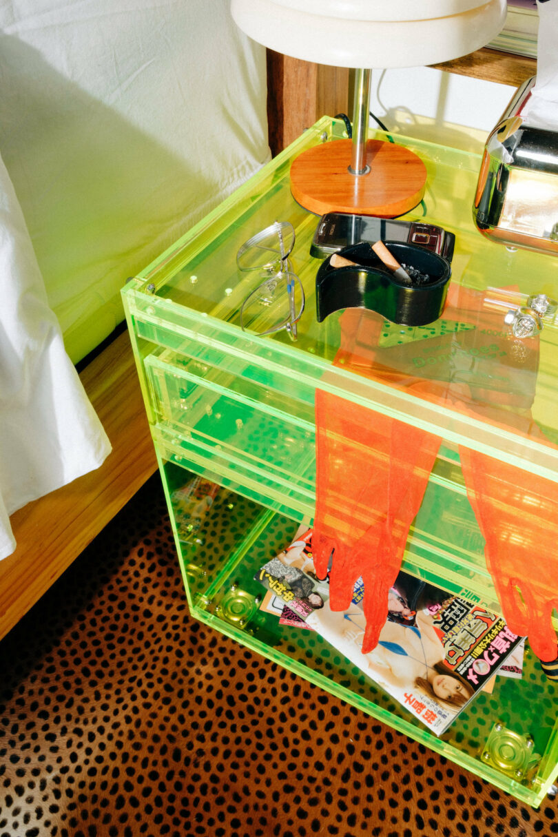 A transparent green bedside table with a lamp, sunglasses, watch, and red gloves. Magazines are on a leopard-print carpet underneath