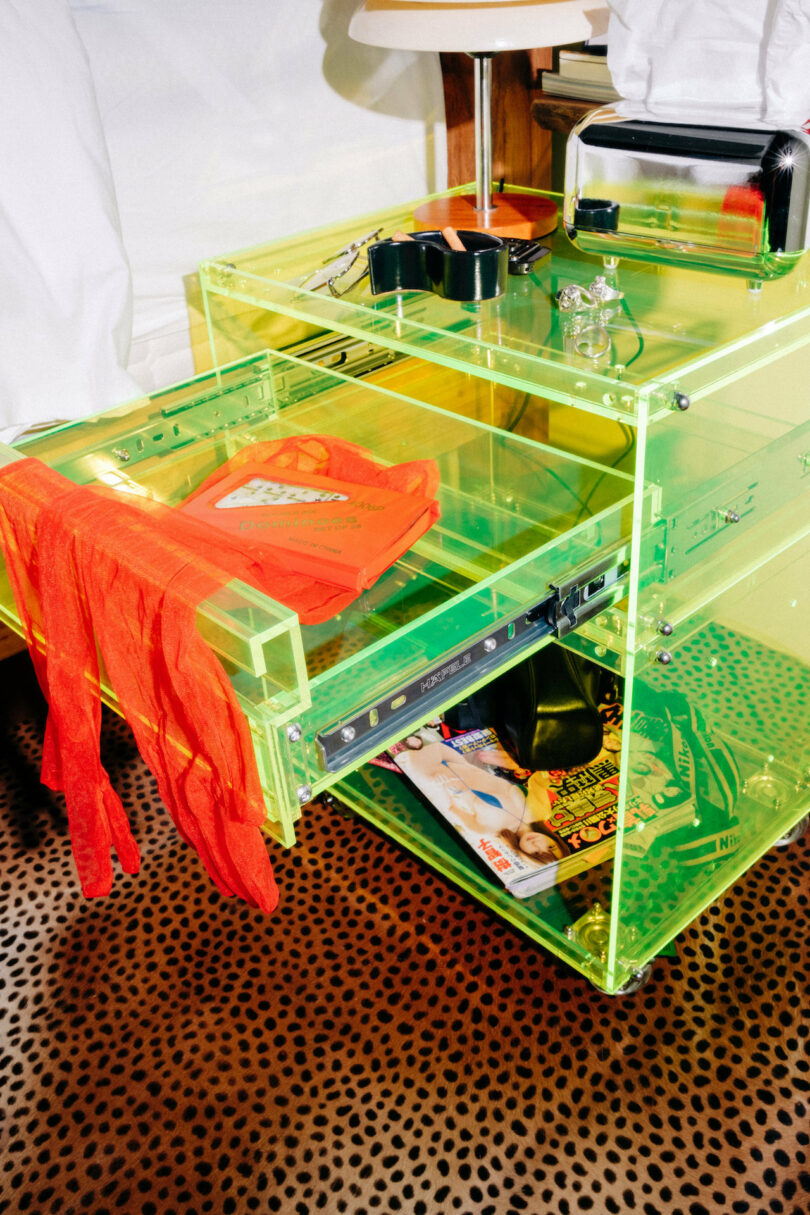 A transparent neon green drawer partly opened contains clothes, magazines, and small items. A red garment hangs from the drawer. The floor is covered with a leopard print rug