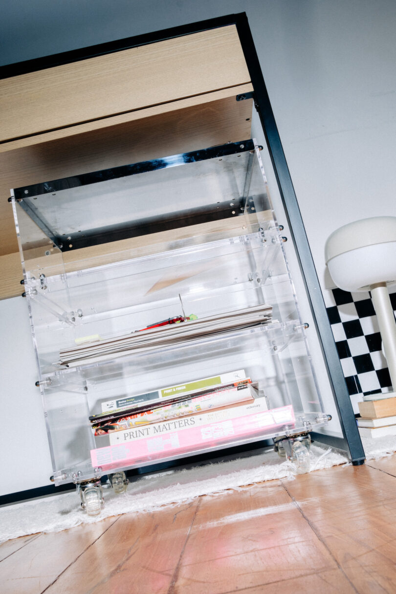 Transparent shelving unit under a wooden desk, holding books and a notepad. A round white stool and checkered wall decor are visible in the background