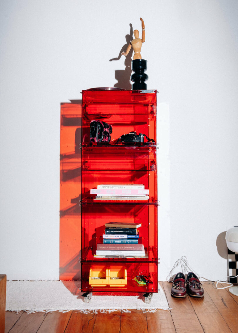 A red translucent shelving unit holds books, decorative items, and shoes. A wooden mannequin poses on top. Cords lay on the floor nearby on wooden flooring