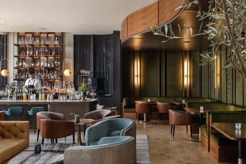 Stylish bar interior with a bartender behind a curved counter. Shelves of bottles are in the background, surrounded by plush green and brown seating areas with dim lighting.