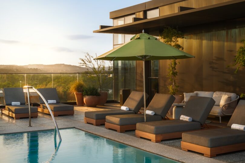 Rooftop pool area with lounge chairs, green umbrella, and potted plants. Glass railing offers a view of the landscape.