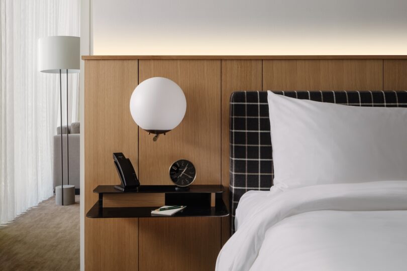 Modern hotel room with a bedside table featuring a phone, clock, and notebook. A spherical lamp is mounted above the table. The bed is neatly made with white linens. A floor lamp is in the background.