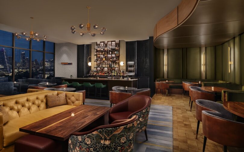 Elegant hotel bar lounge with modern furniture, wooden tables, leather chairs, and a stocked bar. Large windows offer a cityscape view. Ambient lighting creates a cozy atmosphere.