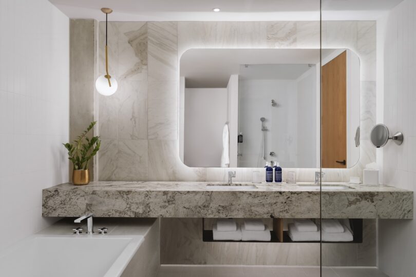 Modern bathroom with a marble vanity, large backlit mirror, bathtub, and potted plant. Wooden accents include a door and shelving with folded towels. A pendant light hangs from the ceiling.