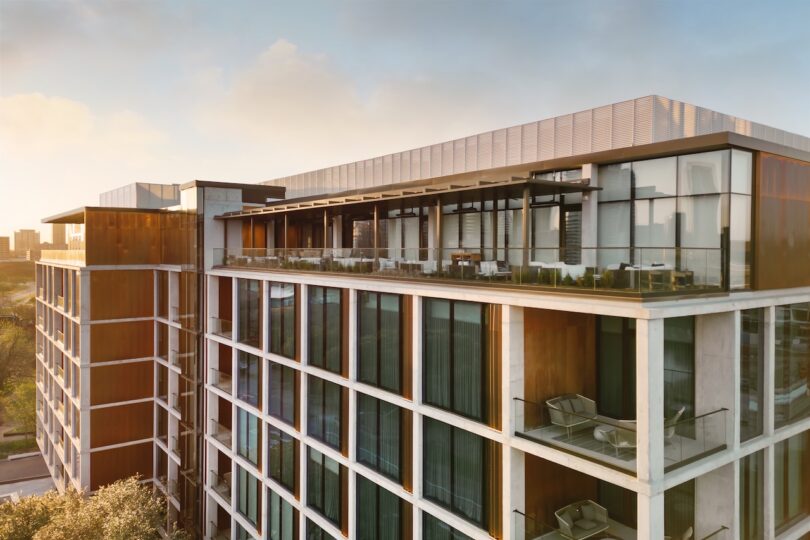 Modern multi-story building with glass façade and balconies, surrounded by trees.