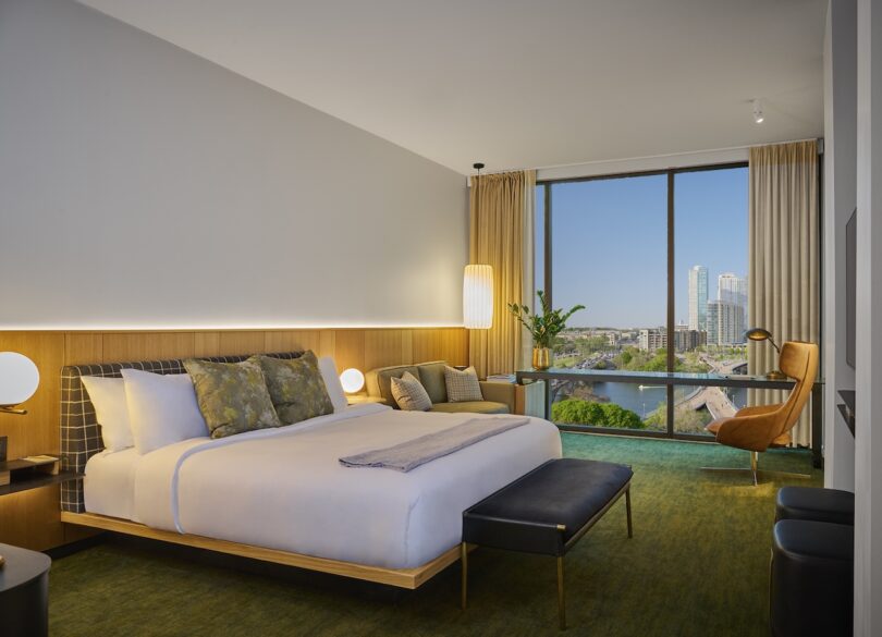 Modern hotel room with a large bed, pillows, and a throw blanket. A desk with a chair is near the window, which offers a cityscape view. Neutral tones and ambient lighting create a cozy atmosphere.