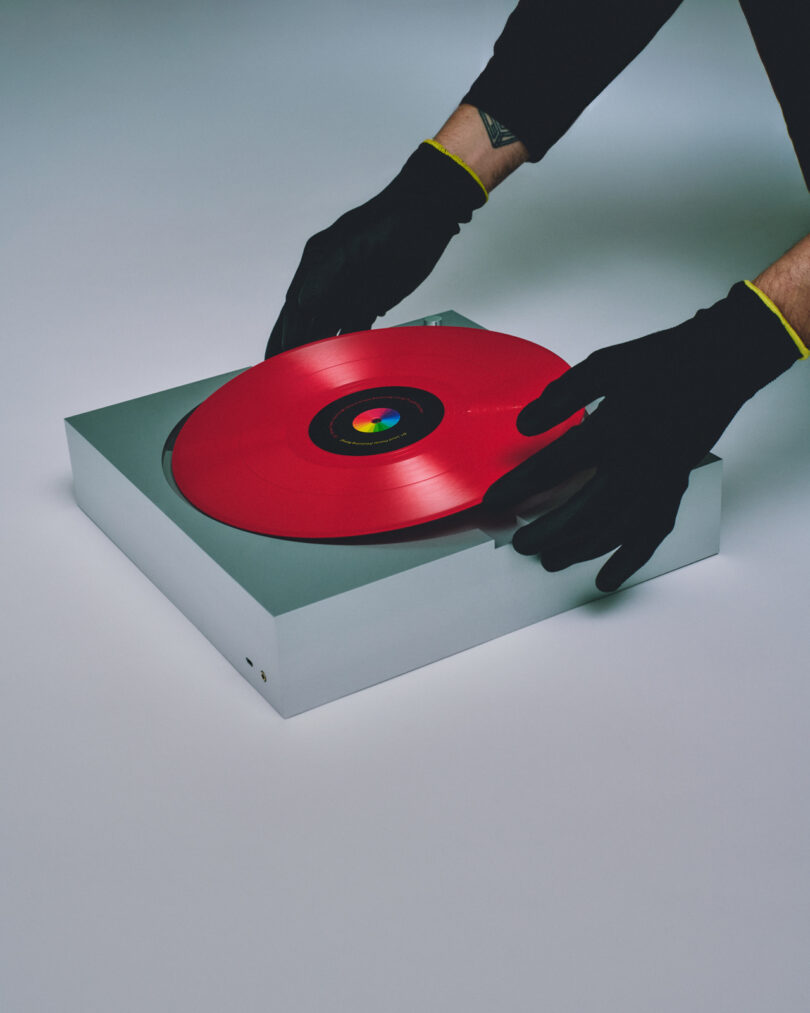 A person wearing black gloves is carefully handling a red vinyl record on a sleek PP-1 white turntable with a gray base.