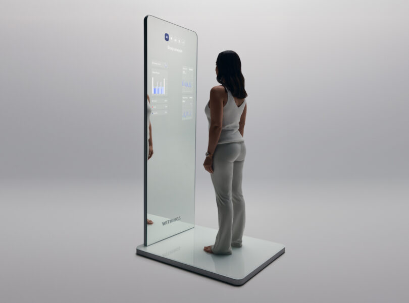 A woman stands on a sleek platform facing a tall smart mirror displaying health data graphs.