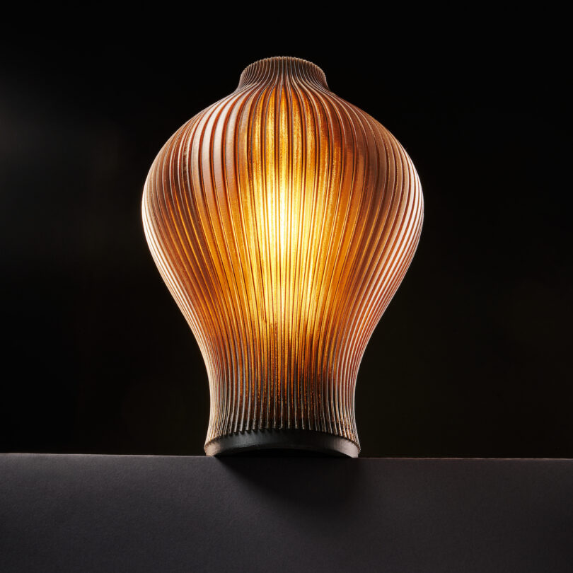 A glowing, ribbed, vase-shaped lamp stands on a dark surface against a black background