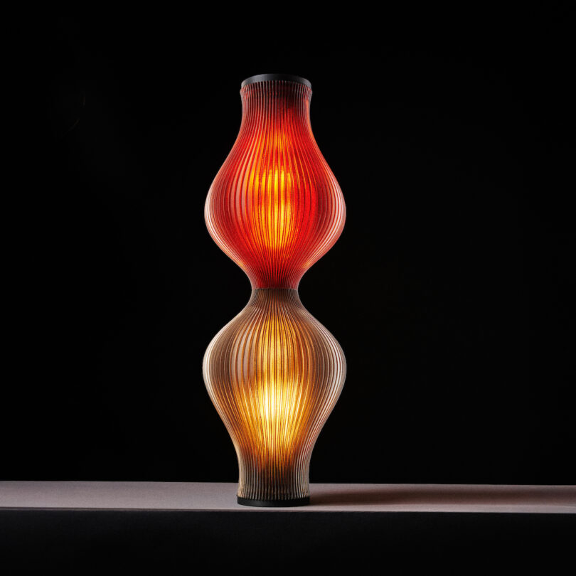 A sculptural lamp with a red upper section and a yellow lower section, both emitting a warm glow. It stands on a flat surface against a black background