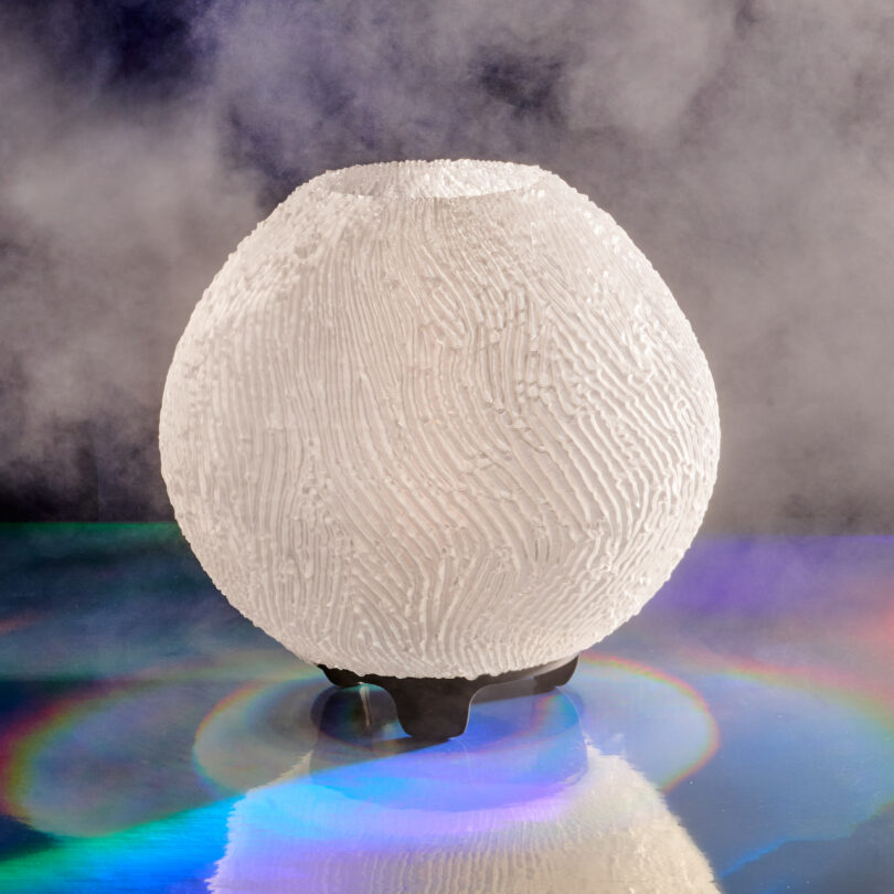 A white, spherical, textured lamp on a reflective surface with mist in the background and circular rainbow light reflections beneath it.