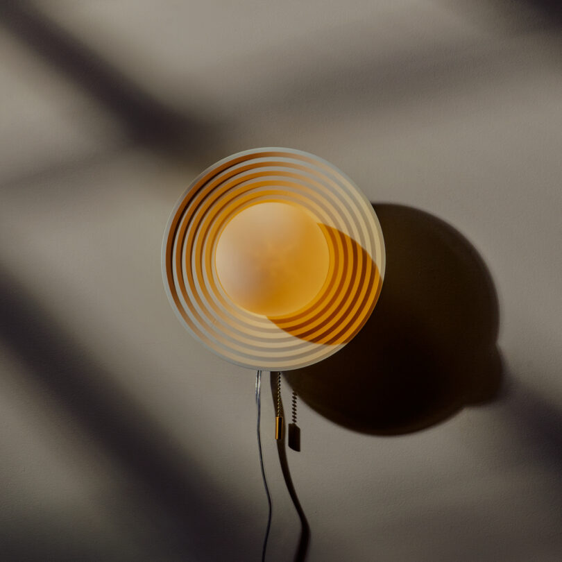 Wall-mounted circular lamp with concentric rings casting a shadow. The lamp emits soft, warm light and has a dangling cord with a switch