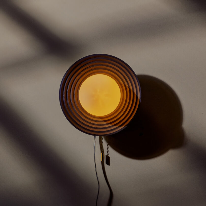 A circular wall lamp emits a warm yellow light, creating a striped shadow pattern on the wall. A cord and pull switch hang below