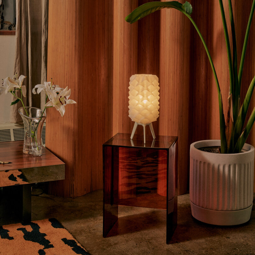 A geometric lamp by Wooj Design sits on a wooden side table next to a potted plant and a vase with white lilies, in a room adorned with wooden walls and a patterned rug