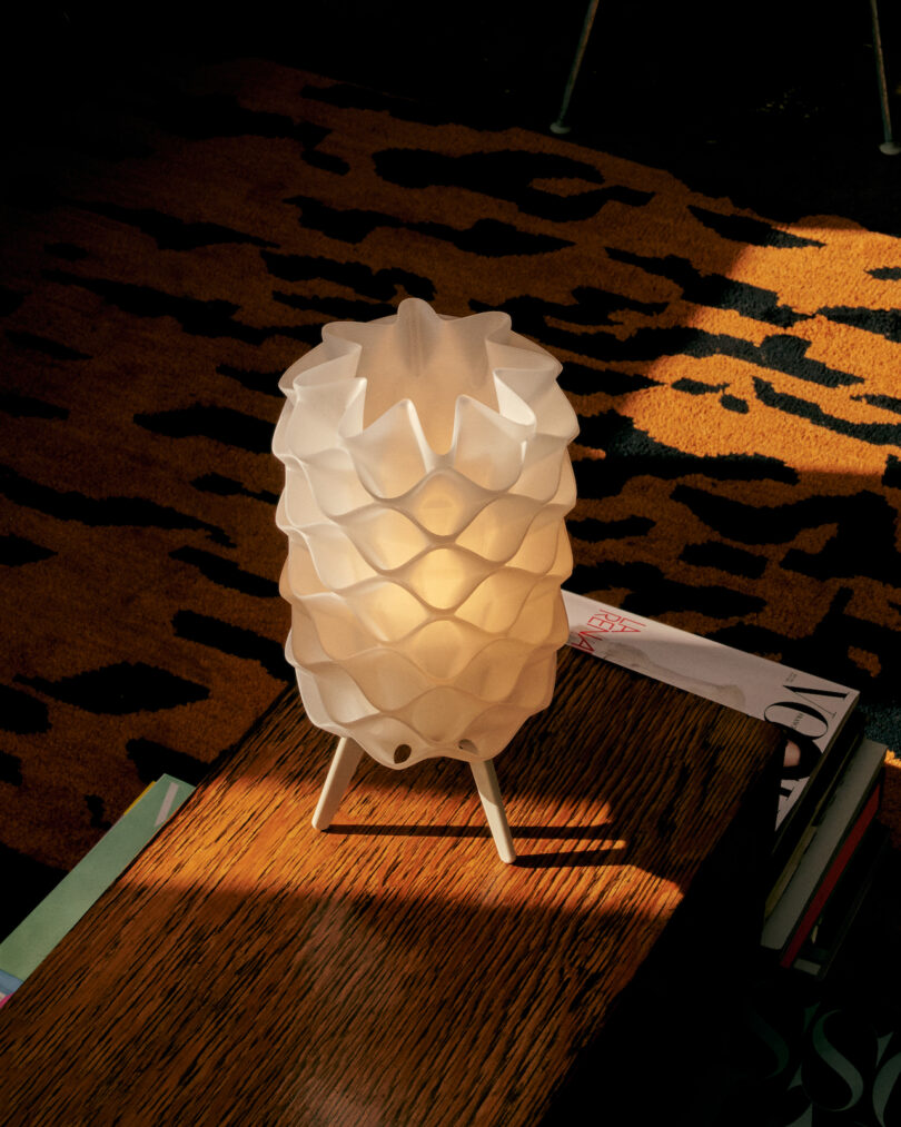 A sculptural white lamp by Wooj Design, showcasing a textured honeycomb pattern, sits elegantly on a wooden surface. The background is adorned with a striking leopard print pattern and partially visible books