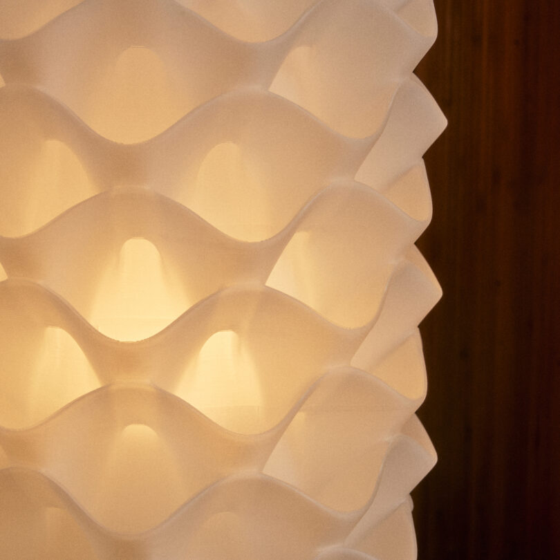 Close-up of a lamp by Wooj Design with a white, wavy, textured pattern. The light is softly diffused through the material, creating a warm glow